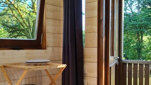 A cozy cabin interior with a wooden shelf holding a kettle, cups, and a plate. A window with a curtain allows natural light in, and a door leads to an outdoor deck.