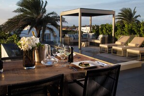 An outdoor dining area with a wooden table set for two, a flower vase, wine glasses, and a plate of food.