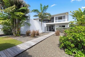 Une maison moderne à deux étages, dotée d’une terrasse en bois menant à l’entrée, entourée d’une végétation luxuriante et baignée par un ciel dégagé.