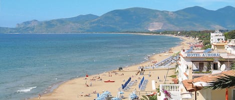 A beach with rows of sunbeds and umbrellas, a lifeguard tower, and a hotel named 'HOTEL AURORA' with a view of the sea and mountains.