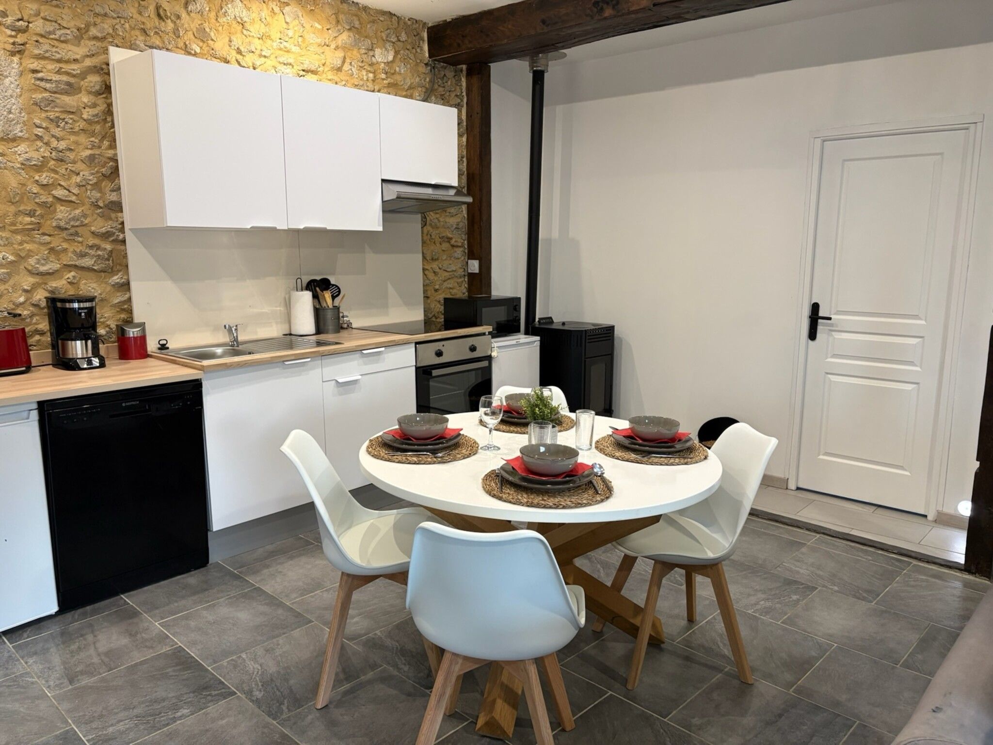 Una cocina moderna con armarios blancos, pared de piedra, una mesa de comedor con platos y vasos, y una cafetera.