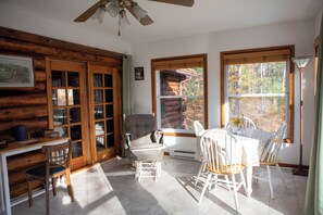 Interior de cabana de tronco com área de jantar, lareira e grandes janelas com vista para a floresta ao redor.