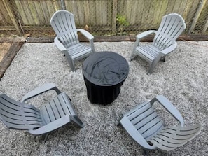 A set of four Adirondack chairs and a round table with a black cover, arranged on a gravel surface.