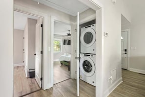 A laundry room with two stacked washing machines, a bed with a green headboard in the adjacent bedroom, and a door leading to another room. - Pet Friendly Family Home Steps to Raleigh Greenway (Raleigh)