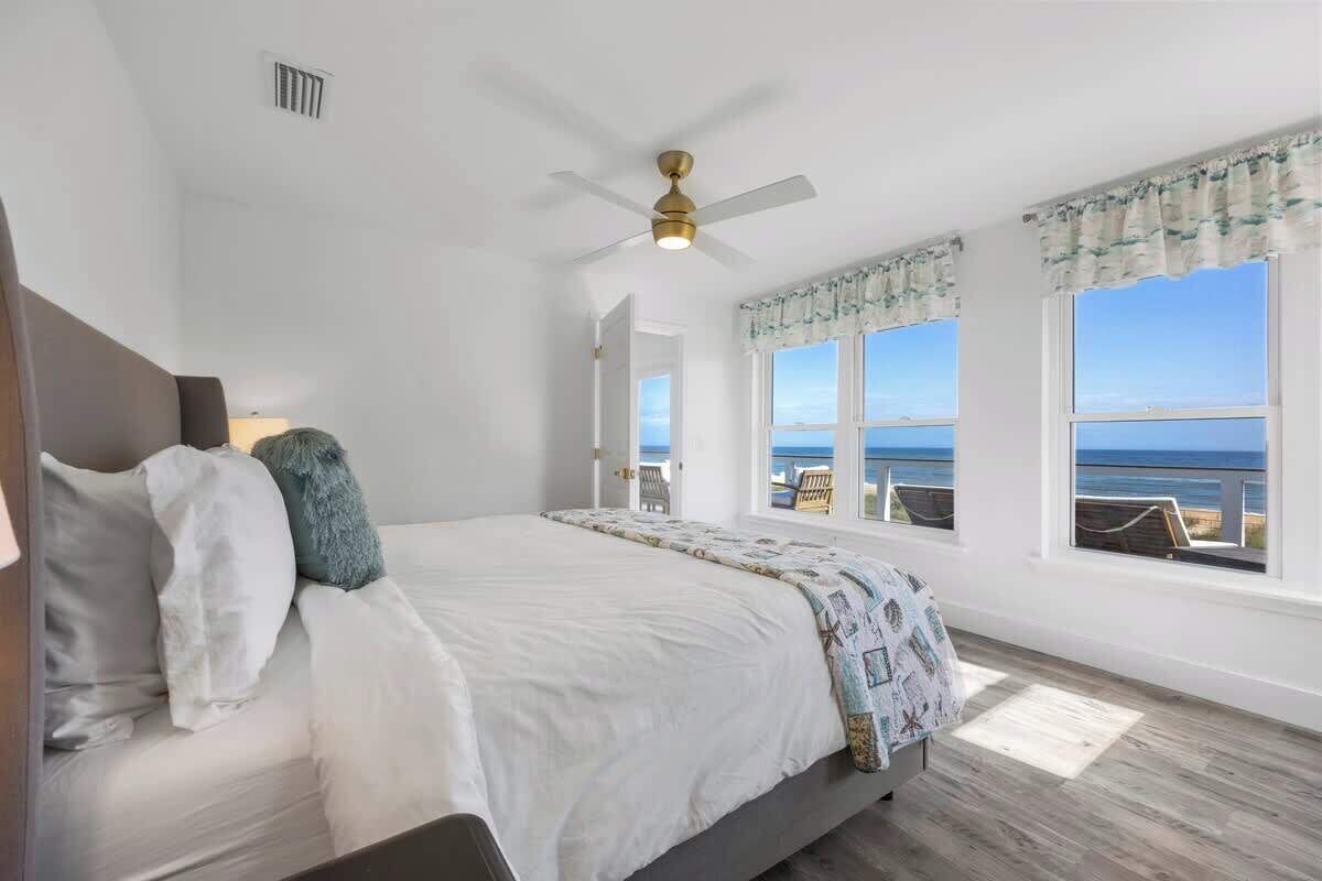 A bedroom with a bed, a ceiling fan, and large windows with a view of the ocean.