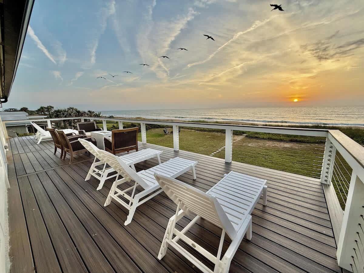 A wooden deck with white lounge chairs and a table, overlooking a sunset over the ocean.