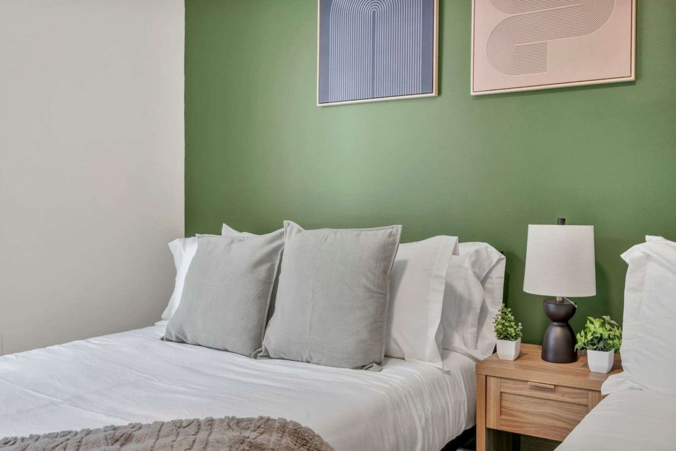 A bedroom with a green wall, a bed with white bedding, a bedside table with a lamp, and two framed artworks.