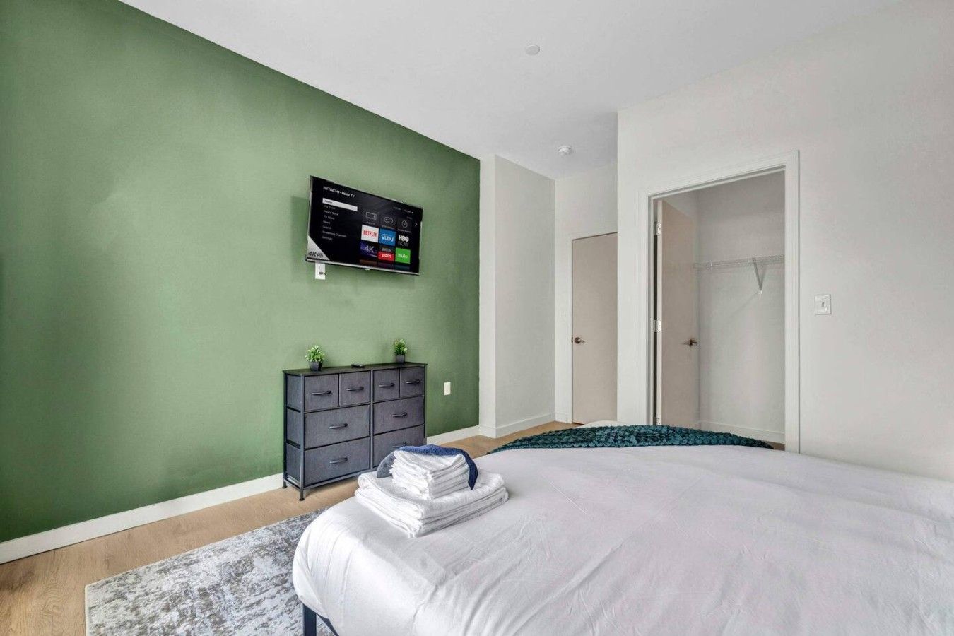 A bedroom with a bed, a dresser, a TV mounted on the wall, and a door leading to another room.