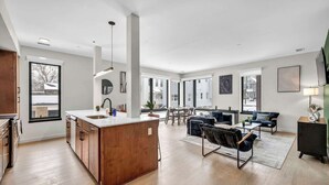 Un vaste espace de vie doté d’un plancher en bois, d’un îlot central avec un évier, d’une table à manger avec des chaises, et d’un coin salon comprenant des canapés et une table basse.