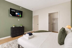 Une chambre à coucher comprenant un lit, une commode, un téléviseur fixé au mur et une porte donnant accès à une salle de bain.