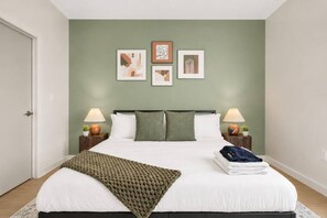 Een net opgemaakt bed met een witte deken en een groene plaid, twee bedlampen en drie ingelijste kunstwerken aan de muur.