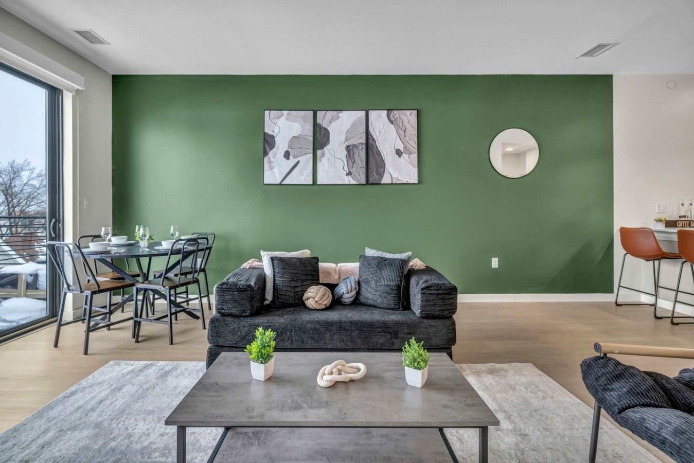 A modern living room with a green accent wall, a dining area with a round table, and a sofa with cushions.