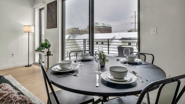 Uma área de refeições com uma mesa redonda posta para quatro pessoas, grandes janelas e vista para edifícios.