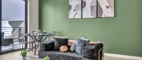 A modern living room with a green accent wall, a gray sofa, a coffee table, and a dining area with a glass door leading to a balcony.