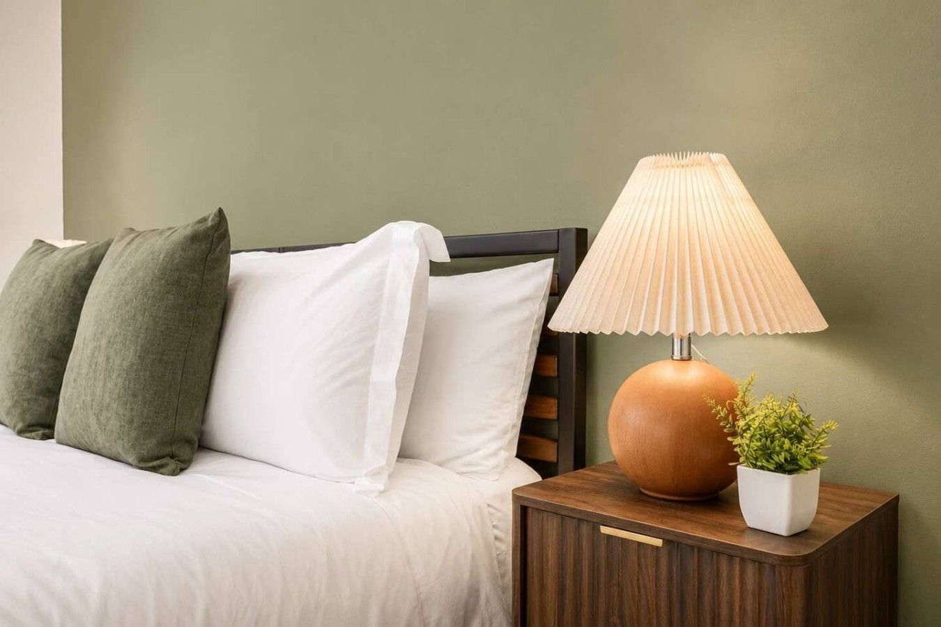 A bed with white and green pillows, a wooden nightstand with a lamp, and a small potted plant.