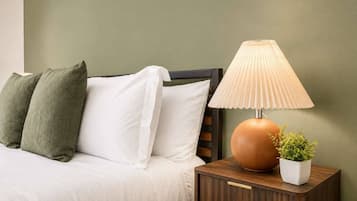 A bed with white and green pillows, a wooden nightstand with a lamp, and a small potted plant.