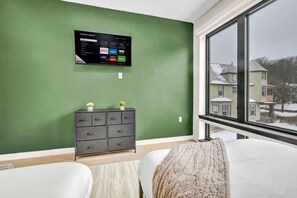 Een slaapkamer met een groene wand, een flatscreen televisie boven een dressoir en een raam met uitzicht op huizen.