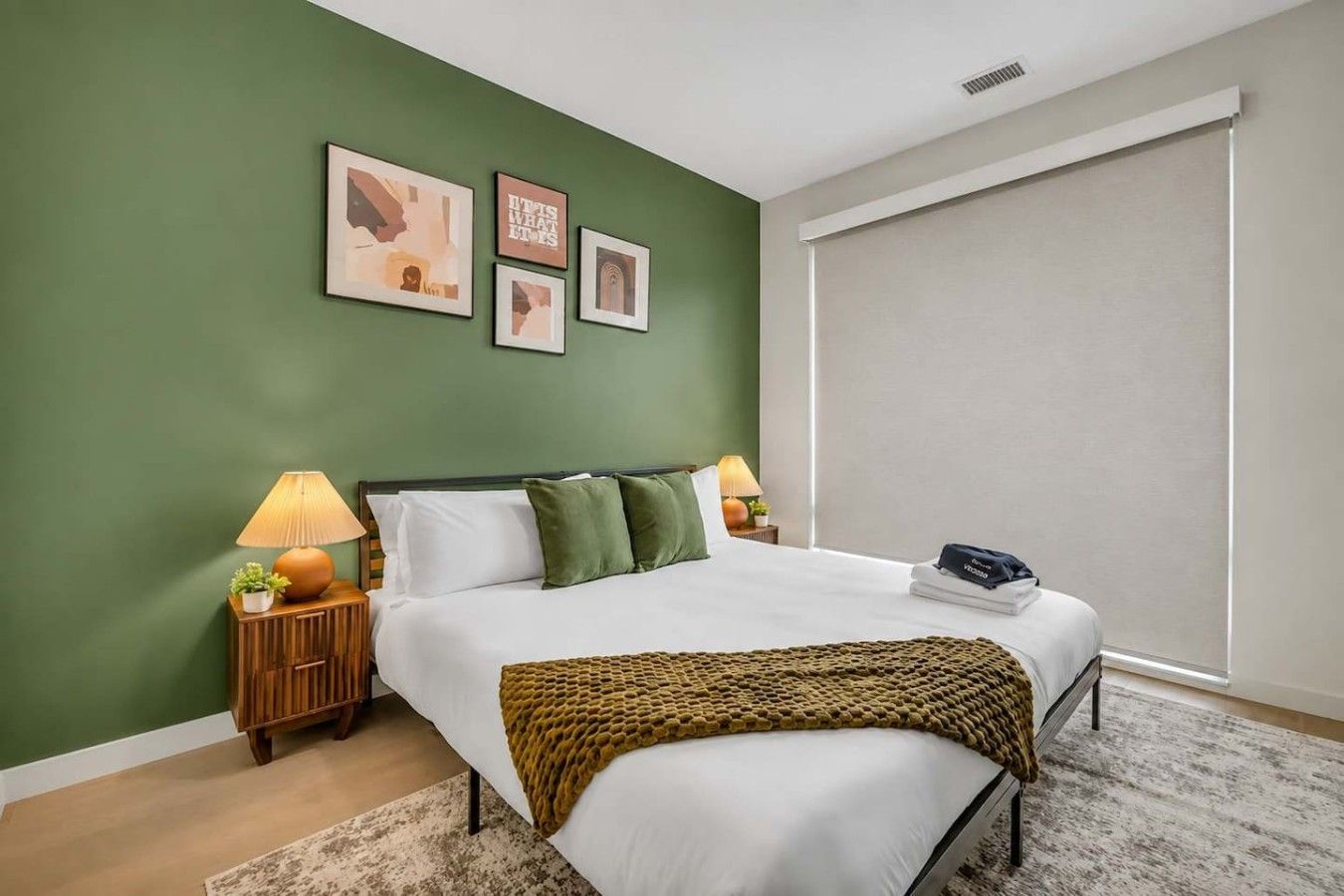 A bedroom with a bed, two bedside tables with lamps, a window with blinds, and three framed pictures on the wall.