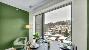 A dining area with a table set for four, a green upholstered chair, and a view of a snowy neighborhood through large windows. - Little Italy & CWRU w King BedGymParkingWD (Cleveland)
