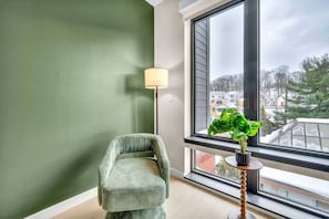 Une pièce avec un mur vert, une fenêtre donnant sur des immeubles, une chaise et un lampadaire.