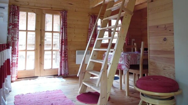 Ein Holzhütten-Interieur mit einer Holzleiter, einem Tisch mit Stühlen und einem großen Fenster mit Vorhängen.
