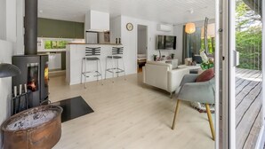 Moderni keittiö, jossa on keskeinen saareke, ruokailutila baarituoleineen, olohuone, jossa on sohva, ja suuri ikkuna, josta avautuu näkymä vehreään maisemaan.