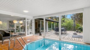 Moderni sisäuimahalli, jossa on läpinäkyvät lasiseinät, puinen terassi ja ulkokalusteilla varustettu patioalue.