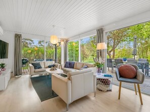 Ein modernes Wohnzimmer mit Sofa, Couchtisch und Sesseln, großen Fenstern mit Blick auf Grünflächen und einer Decke mit weißen Paneelen.