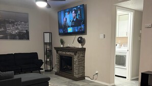 Een woonkamer met een open haard, een flatscreen televisie, een plafondventilator en een groot stadsgezicht schilderij.