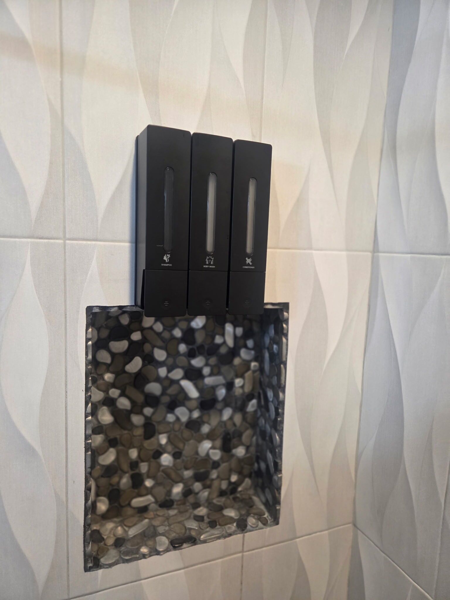 A wall-mounted dispenser with three compartments labeled for shampoo, body wash, and conditioner.
