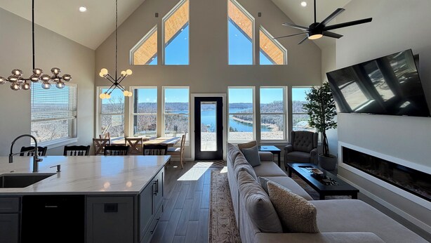 A spacious living area with a high ceiling, large windows, a kitchen with a central island, and a cozy seating area. - Brand-New Beaver Lake Cabin - Lake Views - Fire Pit - Sleeps Up To 9 (Garfield)