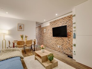 A modern living room with a brick wall, a flat-screen TV, a dining area with wooden chairs, and a sofa. - Atelier Parisien - 4P - Montparnasse Station (Paris)
