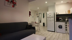 Un espacio habitable compacto con un sofá gris, una mesita blanca, una zona de cocina con lavadora y un refrigerador con imanes.