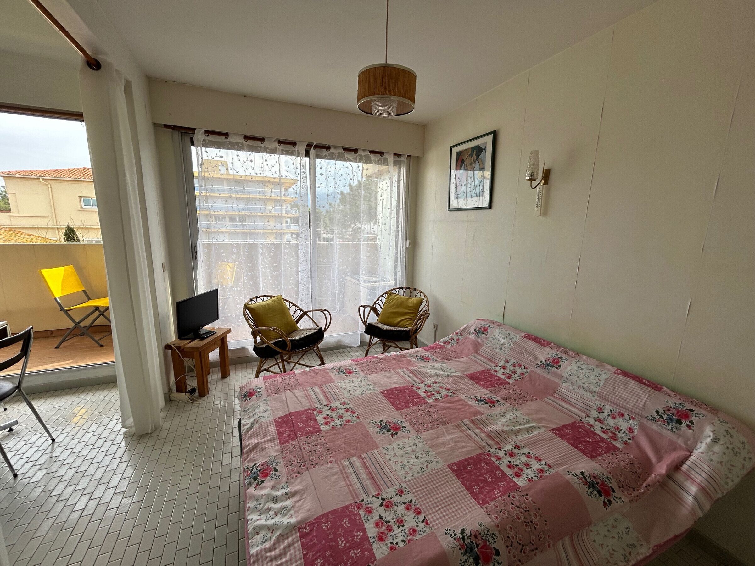 Une chambre à coucher avec un lit, deux chaises, un téléviseur et une fenêtre avec des rideaux.