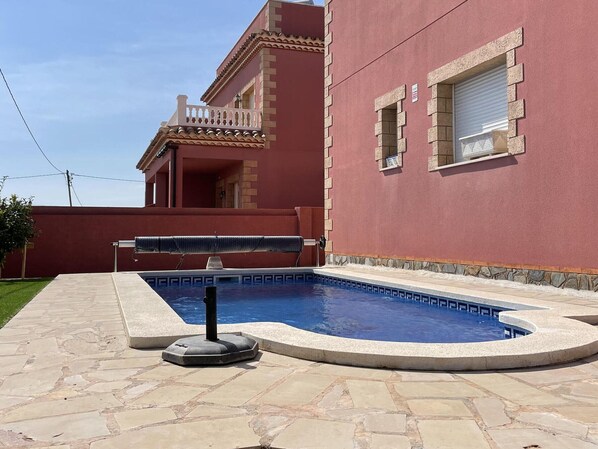 A swimming pool with a stone deck, a red building with a balcony, and a window with a closed shutter.