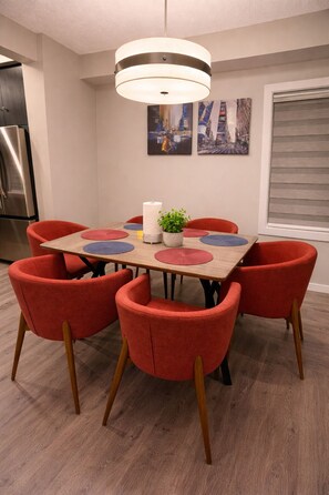A dining area with a wooden table, red upholstered chairs, a hanging light fixture, and two wall-mounted paintings.