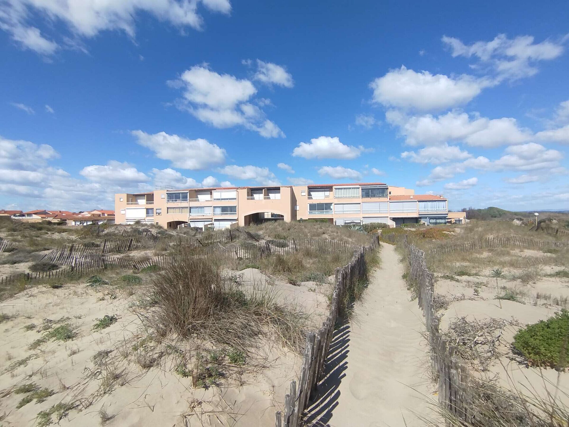 A coastal building with multiple balconies, a sandy path leading to it, and a clear sky.