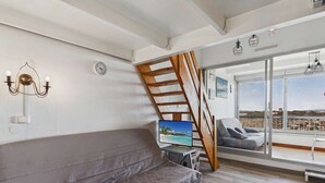 Un salon moderne avec un canapé gris, un téléviseur, un escalier en bois et un balcon offrant une vue dégagée.