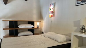 Een eenpersoonsbed met een witte matras en twee kussens, een nachtkastje met een lamp, en een wandlamp.