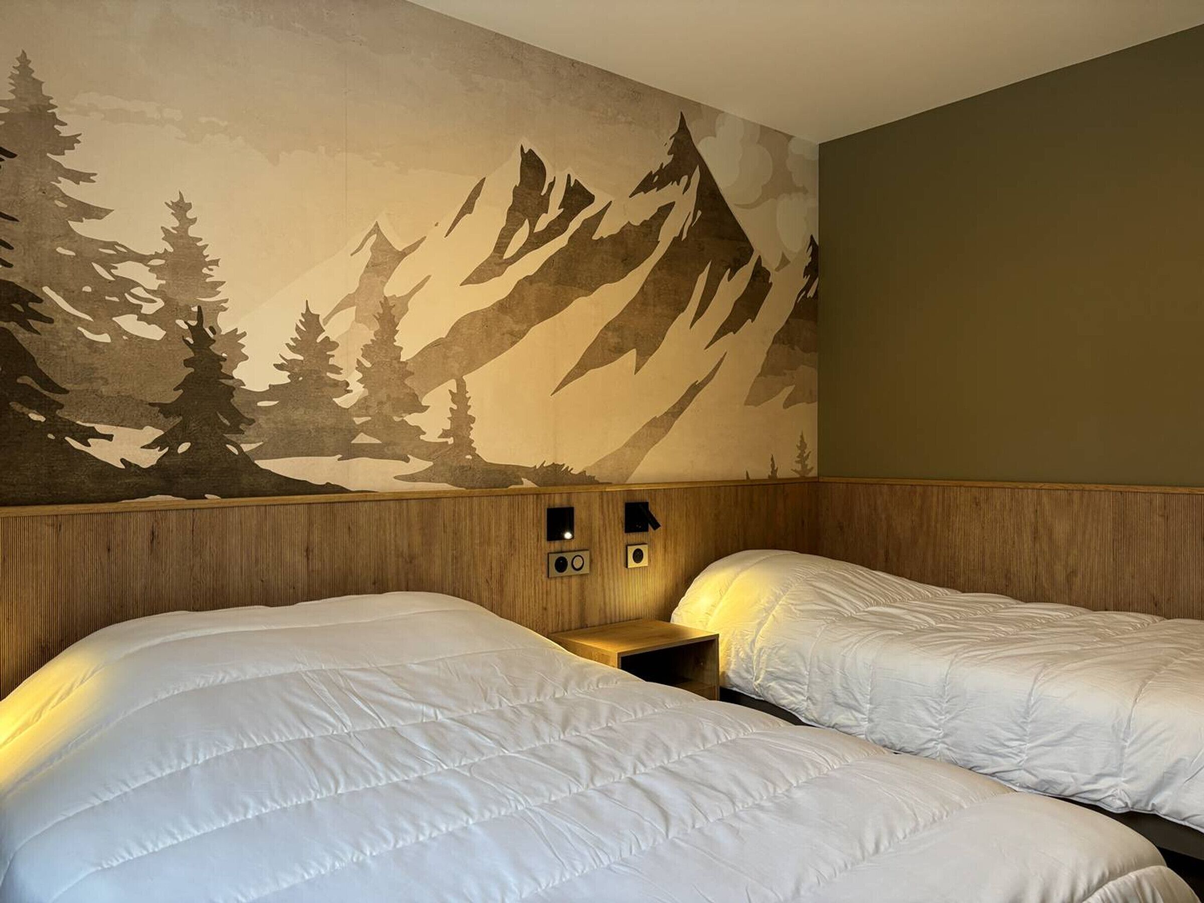 A hotel room with two beds, a wooden headboard, a wall mural of mountains and trees, and a small bedside table with a lamp.