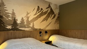 A hotel room with two beds, a wooden headboard, a wall mural of mountains and trees, and a small bedside table with a lamp.