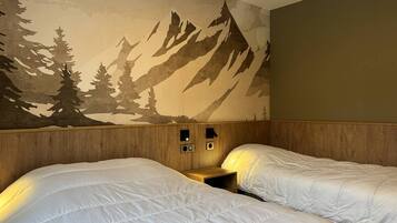 A hotel room with two beds, a wooden headboard, a wall mural of mountains and trees, and a small bedside table with a lamp.