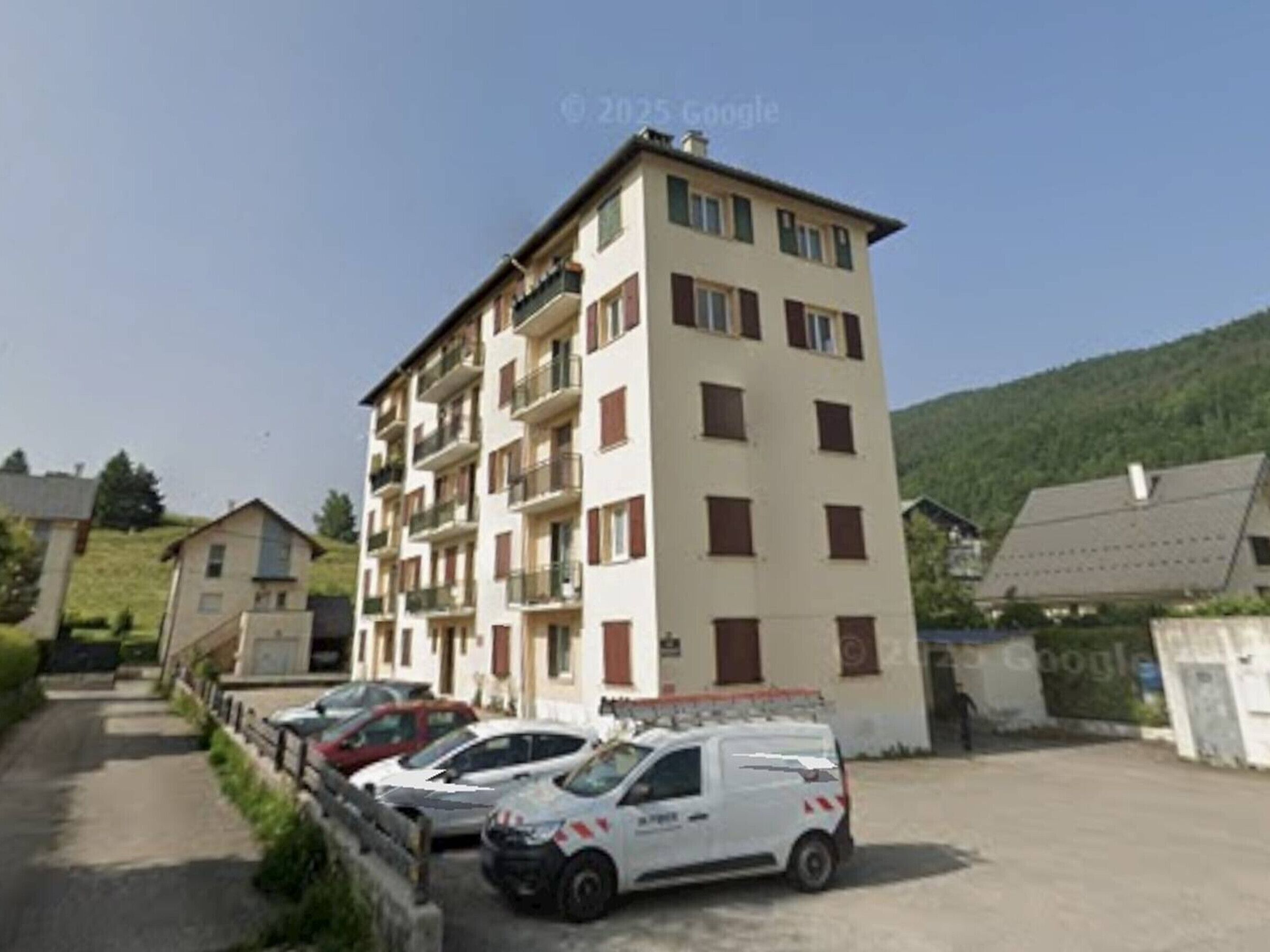A multi-story residential building with balconies, parked cars, and a hilly backdrop.