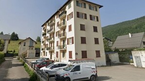 A multi-story residential building with balconies, parked cars, and a hilly backdrop.