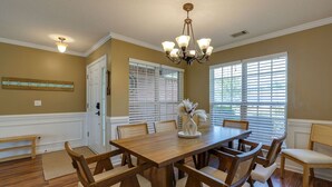 Comedor con mesa y sillas de madera, una lámpara de candelabro y ventanas amplias con persianas.