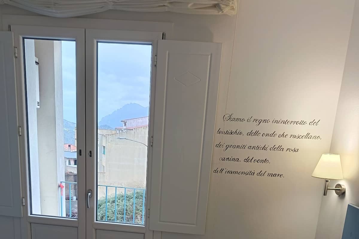 A hotel room with a bed, a nightstand, a wall-mounted lamp, and a view through a door.