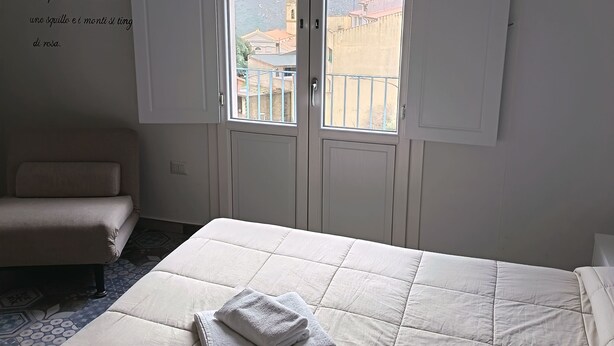 A hotel room with a bed, a sofa, and a window with a view of buildings and mountains. - La Locanda b&b Dandana (Orgosolo)