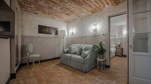 Una stanza con soffitto in mattoni, un divano grigio con cuscini, un tavolino rotondo con una pianta, una sedia, una televisione a schermo piatto e un letto sullo sfondo.