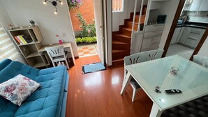 Un espace de vie compact comprenant un canapé bleu, une table à manger en verre, un coin cuisine et un escalier menant à un espace extérieur agrémenté d’un mur en briques et de plantes.