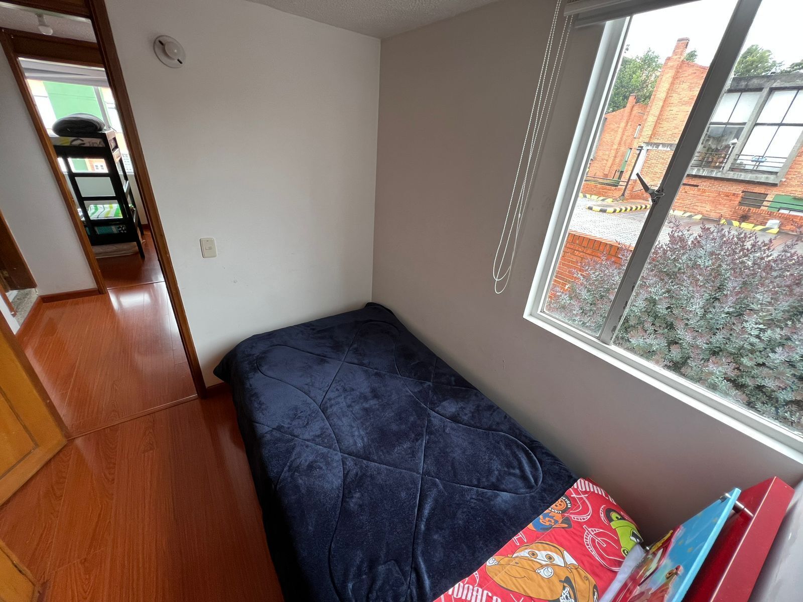 A small, single-bed room with a window overlooking a brick building and greenery.